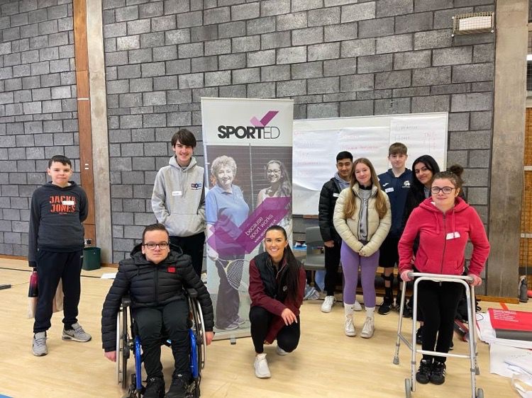 A group of young people infront of a banner with Sported logo. Young people are standing or sitting in wheelchairs or with supports