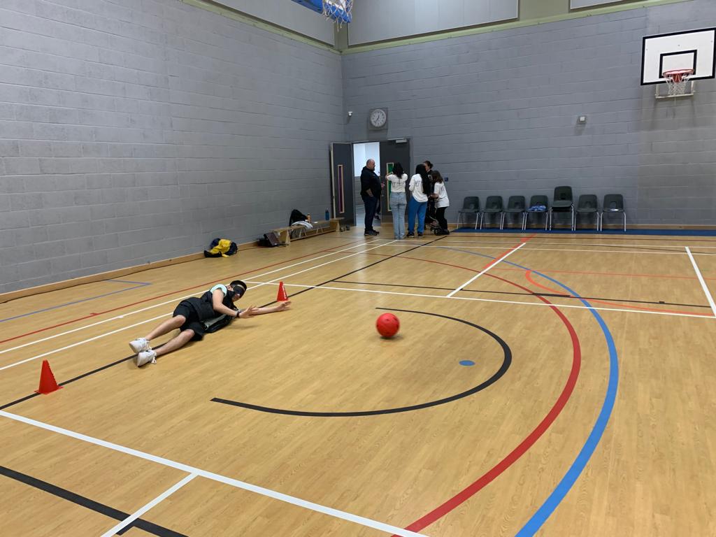 A young person is laying on the floor between two cones blindfolded during a game of goalball. A ball that he has saved from the goal is in front of him