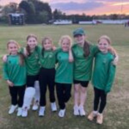 Small group of girls all dressed in green tops with arms round each other looking at the camera. One girls has white cricket pads on her legs.