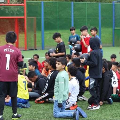 Group of teenagers at a football training session some standing, some kneeling, some sitting all looking like they are listening to the coach