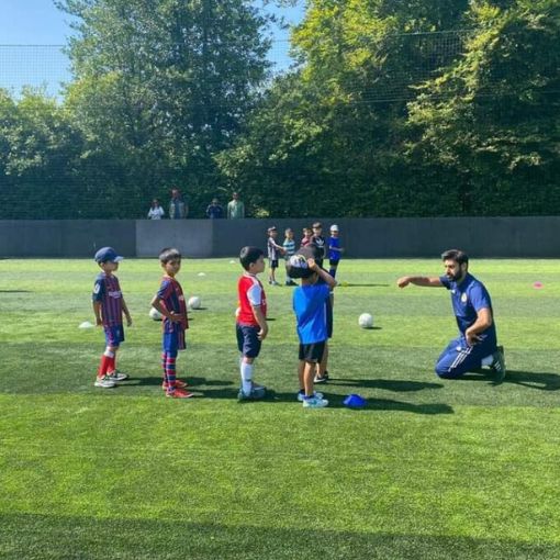 Male coach on his knees in front of a line of four young children on a field of grass with cones and footballs on ground