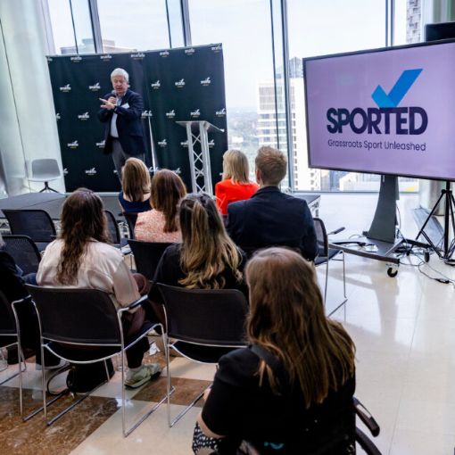 Male presenter speaking with microphone in hand. Audience sitting in chairs looking forward. Screen with the Sported logo.