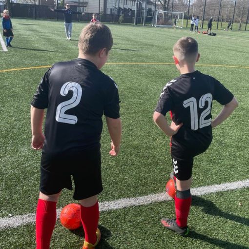 Two boys on football pitch side line with new kits on and footballs at their feet
