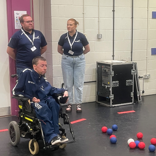 Man in motorised wheelchair with two people stood behind him and Boccia balls on the ground