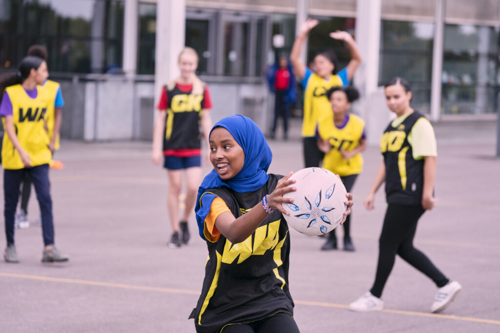 A young girl on a concrete playing surface, holding a ball, smiling. Other children are behind her watching.