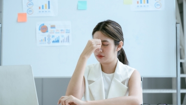 woman, with her hand on the top of her nose, with a whiteboard behind her