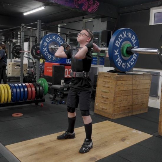 Young person dressed in black holding a heavy weight bar across the front of their chest with puffed out cheeks. In a room with other weight equipment around them.